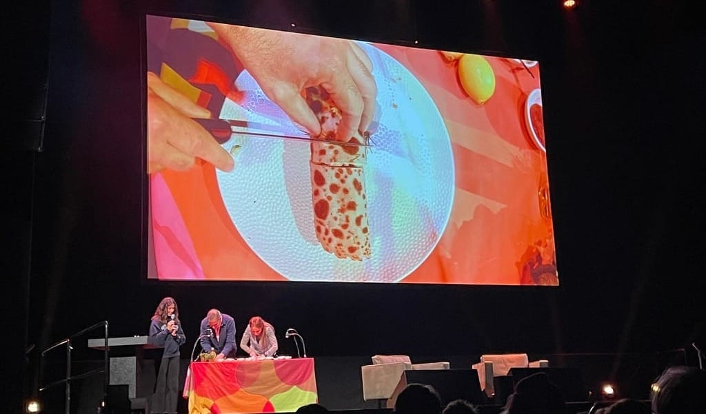 Beeld van podium met twee mensen die pannenkoeken in stukjes snijden.